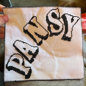 May include: Square, white cross-stitch with the word "NSY" in black, stylized, block letters. The letters have a 3D effect. The edges are finished with a black border. The piece is held by a hand.