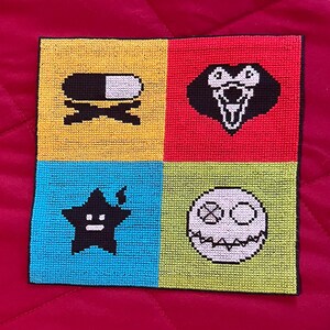May include: A square, cross-stitch piece with four coloured quadrants: yellow, red, blue, and green. Each quadrant features a black and white graphic: a pill, a face, a star, and a skull. The piece is likely handmade.