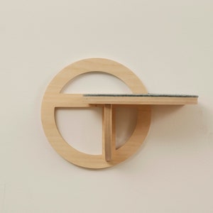 May include: A modern, wall-mounted cat shelf made of light-colored wood. The design features a circular element with a rectangular shelf and a supporting structure. The shelf has a dark gray, textured surface, providing a comfortable spot for a cat.