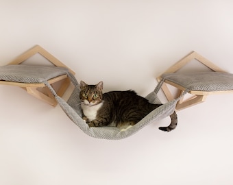 May include: A gray cat hammock with two wooden platforms attached to a wall. The hammock is made of a soft, gray fabric and has a gray cushion on each platform. A tabby cat is lying in the hammock.