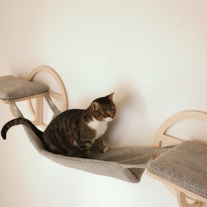 Op de afbeelding: Een kat zit in een hangmat-achtig kattenbed dat is bevestigd aan een aan de muur gemonteerd houten plank. De plank heeft een grijs kussen en een rond houten frame. Rechts van de hangmat is er een tweede plank met een grijs kussen en een rond houten frame.