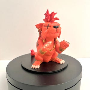 May include: A small, orange, anthropomorphic creature figurine with spiky hair, a black cross marking over one eye, and gold-colored bands on its arms and neck. The figure has white claws and black Japanese characters printed on its right shoulder.