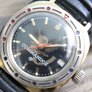 May include: A black and gold wristwatch with a black leather strap. The watch face features a gold eagle with the word "Commandos" printed below it. The watch has a red second hand and a date window at the 4 o'clock position.