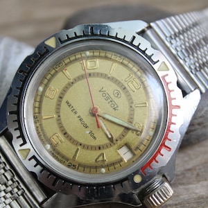 May include: A vintage Vostok watch with a gold-coloured face, black numerals, and a silver metal strap. The watch face has the text "Vostok WATER PROOF 20m". The bezel has red and white accents.