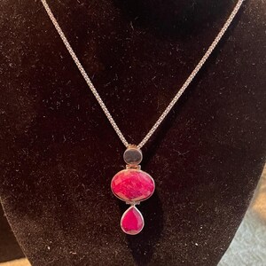 May include: A silver chain necklace with a pendant featuring three red gemstones. The pendant is made up of a round gemstone, a larger oval gemstone, and a teardrop-shaped gemstone.