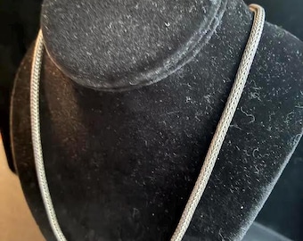 May include: A silver-colored necklace with a woven chain design is displayed on a black velvet bust. The necklace is draped around the bust, showcasing its intricate pattern and metallic sheen. The background is dark, emphasizing the jewelry.