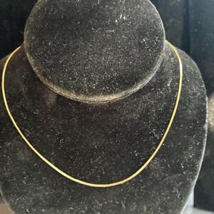 May include: A delicate gold-colored chain necklace displayed on a black velvet jewelry bust. The chain has a fine, textured design and is presented against a dark background, highlighting its subtle shine. This necklace is a classic jewelry piece.