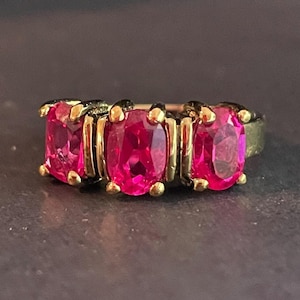 May include: A gold-toned ring featuring three oval-cut, vibrant pink gemstones. The gemstones are set in individual gold bezels, with small gold accents on the sides. The ring's band has a dark, possibly oxidized, finish.