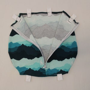May include: A round, zippered bag with a mountain range pattern in shades of teal, blue, and gray. The bag has white tabs with snaps around the top edge. The zipper is white.