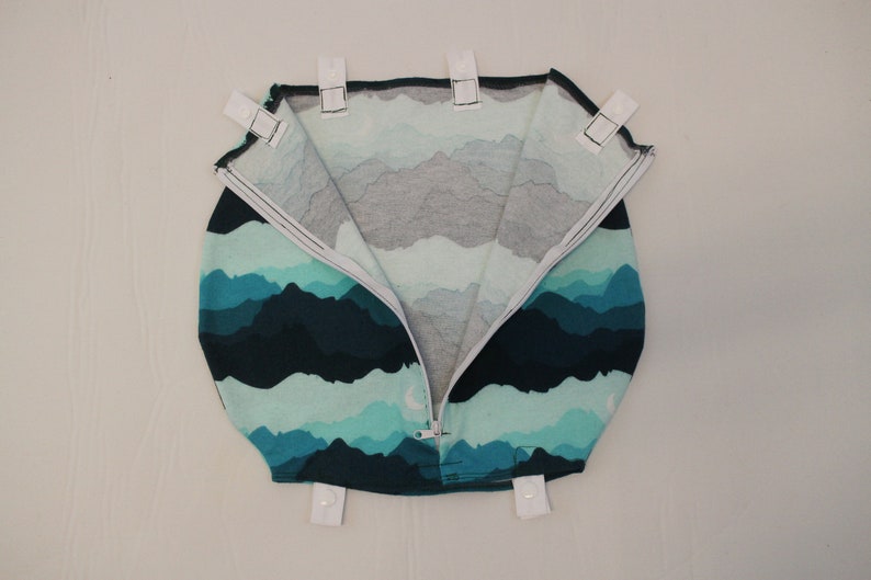May include: A round, zippered bag with a mountain range pattern in shades of teal, blue, and gray. The bag has white tabs with snaps around the top edge. The zipper is white.