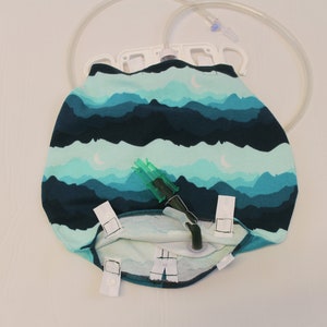 May include: A medical drainage bag with a mountain and moon pattern in shades of teal and navy blue. The bag has a clear plastic tube and a green connector. White clips secure the bag.