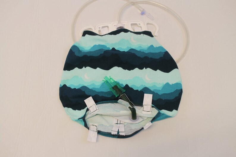 May include: A medical drainage bag with a mountain and moon pattern in shades of teal and navy blue. The bag has a clear plastic tube and a green connector. White clips secure the bag.
