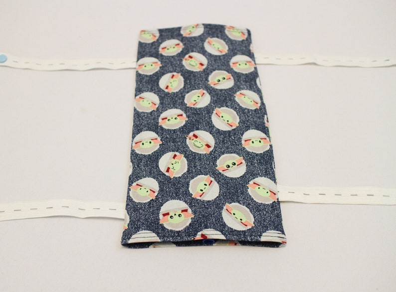 May include: A rectangular, dark blue fabric item with a repeating pattern of green-skinned, large-eyed characters in white circles. White elastic bands extend from the sides. The item appears to be a case or cover.