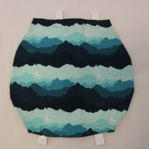 May include: A fabric item with a mountain range design in shades of teal and navy blue. The design includes crescent moons. White snaps are attached at the top and bottom. The item is likely a baby product.