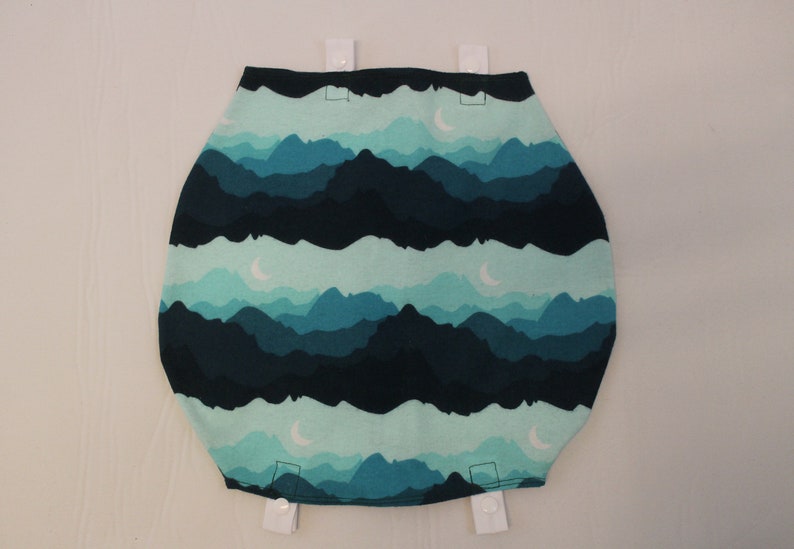 May include: A fabric item with a mountain range design in shades of teal and navy blue. The design includes crescent moons. White snaps are attached at the top and bottom. The item is likely a baby product.