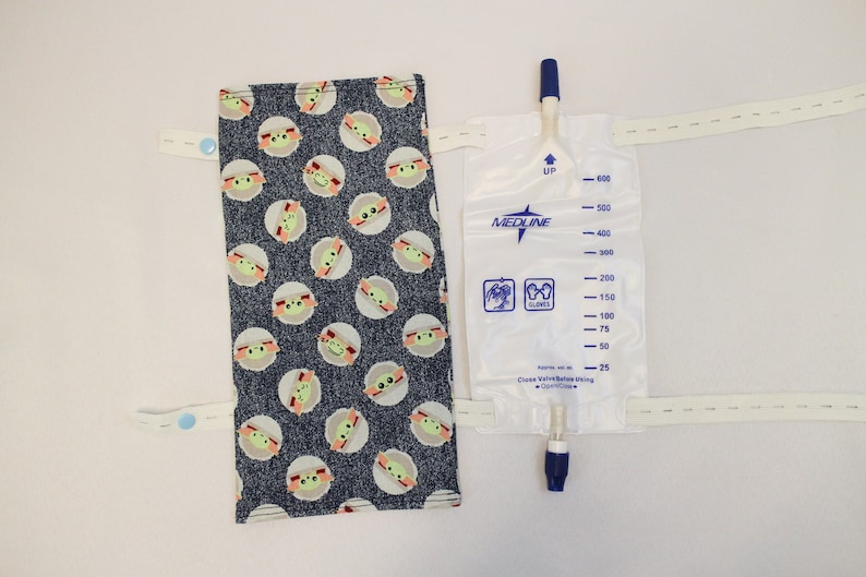 May include: A medical urine bag with a blue and white design, featuring a fabric cover with a repeating pattern of a cartoon character. The bag has measurement markings and a blue valve.