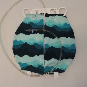 May include: A medical bag with a mountain landscape pattern in shades of teal and navy blue. The bag has a zipper closure and a clear tube attached. The bag is suspended from a white plastic hanger.