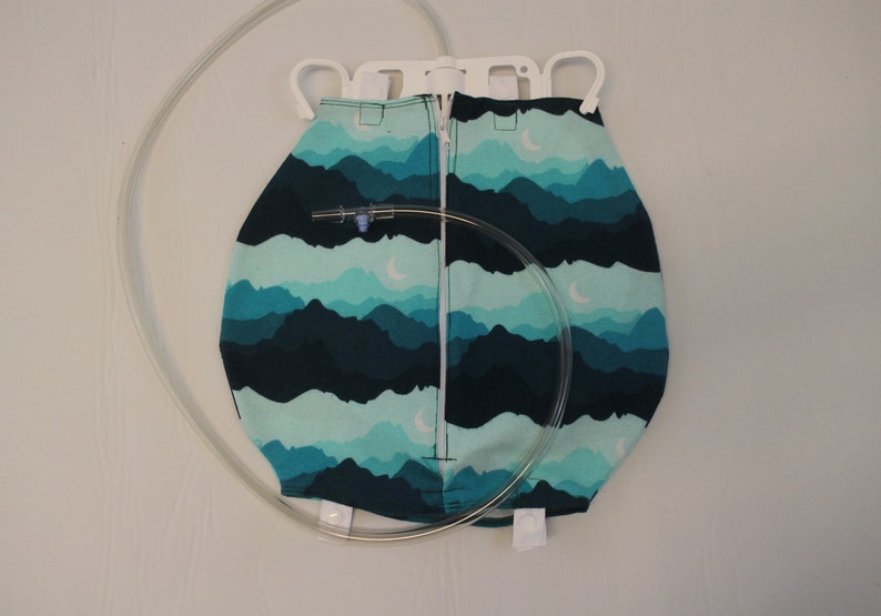 May include: A medical bag with a mountain landscape pattern in shades of teal and navy blue. The bag has a zipper closure and a clear tube attached. The bag is suspended from a white plastic hanger.