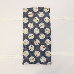 May include: A rectangular fabric item with a dark blue background and a pattern of light green and tan cartoon character faces. Two white elastic straps with blue buttons are attached.