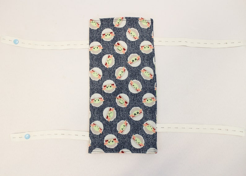 May include: A rectangular fabric item with a dark blue background and a pattern of light green and tan cartoon character faces. Two white elastic straps with blue buttons are attached.