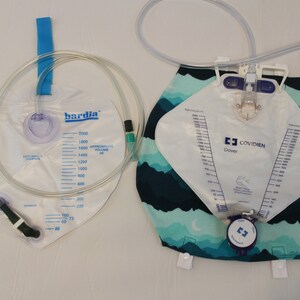 May include: Two clear plastic medical urine collection bags. One bag is labeled "bardia" and has a blue strap. The other bag is labeled "COVIDIEN Dover" and has a patterned fabric backing with blue and green colors. Both bags have measurement markings.