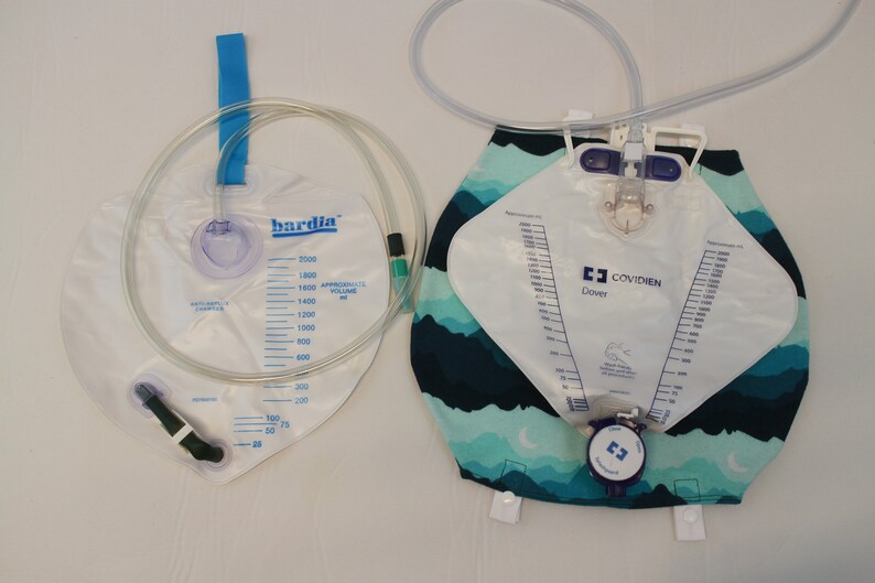 May include: Two clear plastic medical urine collection bags. One bag is labeled "bardia" and has a blue strap. The other bag is labeled "COVIDIEN Dover" and has a patterned fabric backing with blue and green colors. Both bags have measurement markings.
