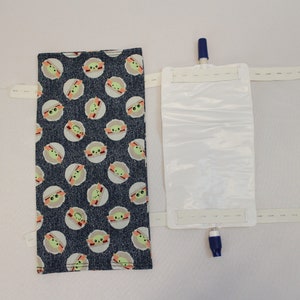 May include: A medical device with a blue and white plastic bag and a fabric cover. The fabric cover is dark blue with a pattern of green and white cartoon character faces. The device has white straps with velcro closures.