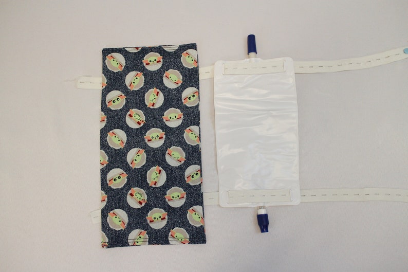 May include: A medical device with a blue and white plastic bag and a fabric cover. The fabric cover is dark blue with a pattern of green and white cartoon character faces. The device has white straps with velcro closures.