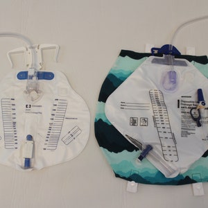 May include: Two disposable urine drainage bags. One is white with a clear tube and a scale printed on the front. The other is white with a blue and green patterned backing and a clear tube. Both have measurement scales.