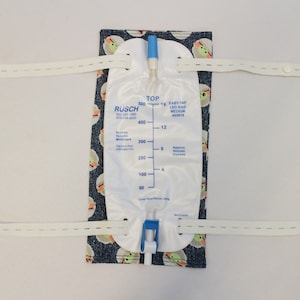 May include: A white leg bag with a blue top and a blue valve, attached to a patterned fabric with a cartoon character design. The bag has volume markings in milliliters and ounces. White straps are attached to the fabric.