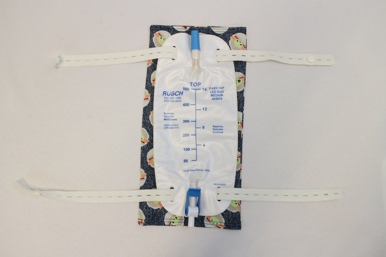 May include: A white leg bag with a blue top and a blue valve, attached to a patterned fabric with a cartoon character design. The bag has volume markings in milliliters and ounces. White straps are attached to the fabric.