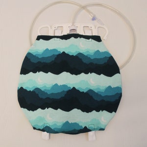 May include: A medical IV bag cover with a mountain and moon pattern in shades of teal and navy blue. The cover is designed to fit over an IV bag, with a white plastic hanger and clear tubing visible at the top.