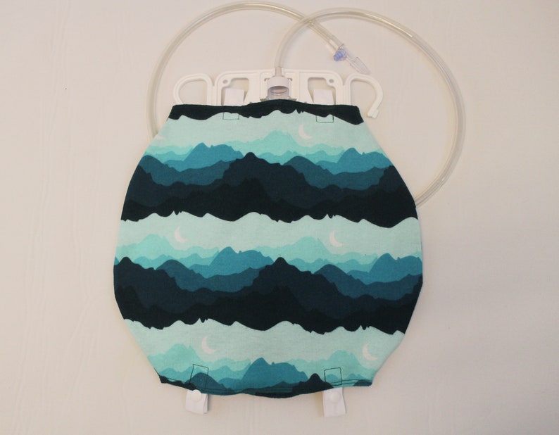 May include: A medical IV bag cover with a mountain and moon pattern in shades of teal and navy blue. The cover is designed to fit over an IV bag, with a white plastic hanger and clear tubing visible at the top.