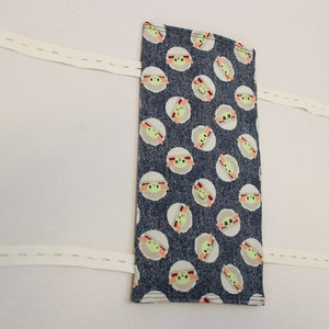 May include: A rectangular fabric item with a dark blue background and a repeating pattern of a cartoon character's face. Attached are three white elastic bands with blue buttons. The item is likely a face mask or similar accessory.