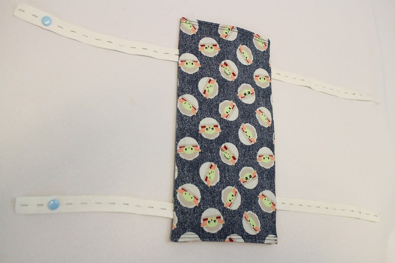May include: A rectangular fabric item with a dark blue background and a repeating pattern of a cartoon character's face. Attached are three white elastic bands with blue buttons. The item is likely a face mask or similar accessory.
