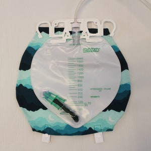 May include: A clear Bard brand urine collection bag with a capacity of 2000 ml, featuring a green and white measurement scale. The bag is attached to a fabric cover with a blue and teal mountain range pattern. A clear tube extends from the top.