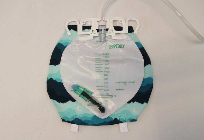 May include: A clear Bard brand urine collection bag with a capacity of 2000 ml, featuring a green and white measurement scale. The bag is attached to a fabric cover with a blue and teal mountain range pattern. A clear tube extends from the top.