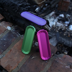 May include: Three small, rectangular metal trays in purple, green, and pink. The trays are sitting on a brick surface with a charred wooden log behind them.