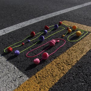 May include: Colorful, abstract necklaces with various shapes and sizes of beads. The beads are red, purple, pink, and yellow, strung on neon green and pink cords. The necklaces are arranged on a dark asphalt surface with white and yellow painted lines.