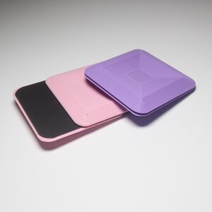 K&ouml;nnte beinhalten: Drei quadratische Untersetzer, einer lila, einer rosa und einer schwarz. Die lila und rosa Untersetzer haben ein strukturiertes, geschichtetes Design.
