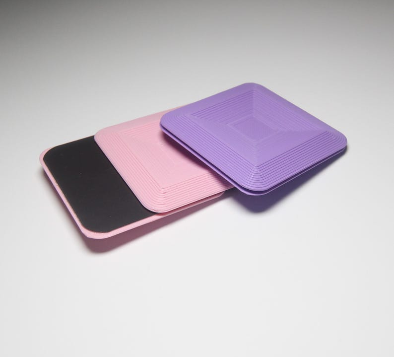 K&ouml;nnte beinhalten: Drei quadratische Untersetzer, einer lila, einer rosa und einer schwarz. Die lila und rosa Untersetzer haben ein strukturiertes, geschichtetes Design.
