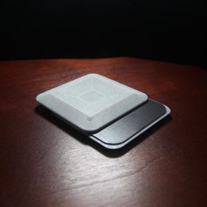 May include: A white, square-shaped, 3D printed object with a concentric circle pattern on top. The object is layered on top of a black, rectangular, magnetic base.