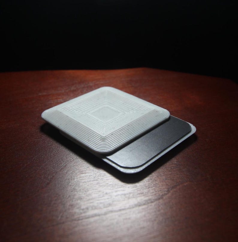 K&ouml;nnte beinhalten: Ein wei&szlig;es, quadratisches, 3D-gedrucktes Objekt mit einem konzentrischen Kreis-Muster auf der Oberseite. Das Objekt ist auf einer schwarzen, rechteckigen, magnetischen Basis geschichtet.