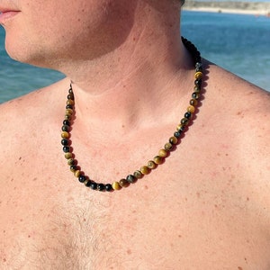 May include: A man wearing sunglasses and a necklace made of tiger's eye beads. He is standing on a beach with the ocean in the background.