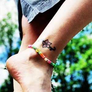 Puede incluir: Un tobillo de mujer con un tobillo de cuentas de colores y un pequeño tatuaje de estrella. El tobillo tiene un amuleto de cara sonriente.