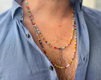 Op de afbeelding: Een meerkleurige kralenketting met een gelaagd ontwerp. De ketting is gemaakt van kleine, kleurrijke kralen en wordt gedragen door een persoon met een blauw gestreept overhemd.