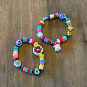 May include: Two colorful beaded bracelets with various charms, including smiley faces, flowers, and a mushroom. The bracelets are made of bright colors like pink, yellow, green, blue, and purple.