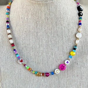 Op de afbeelding: Een kleurrijke kralenketting met verschillende bedels, waaronder harten, smiley-gezichten en een yin-yang symbool. De ketting is gemaakt met witte parels, gouden kralen en gekleurde glazen kralen.