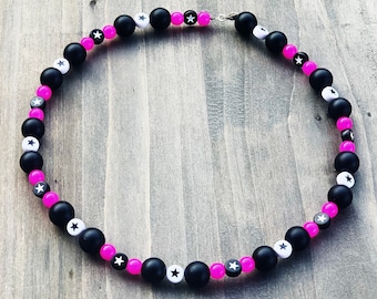 May include: A black and pink beaded necklace with white stars. The necklace is made of round beads with a matte finish.