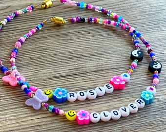 Op de afbeelding: Twee kleurrijke kralenkettingen met de namen "Rosie" en "Clover" gespeld in witte kralen. De kettingen hebben roze, blauwe en gele bloemen, vlinders en smiley-gezichten. De kettingen zijn verbonden met gouden sluitingen.